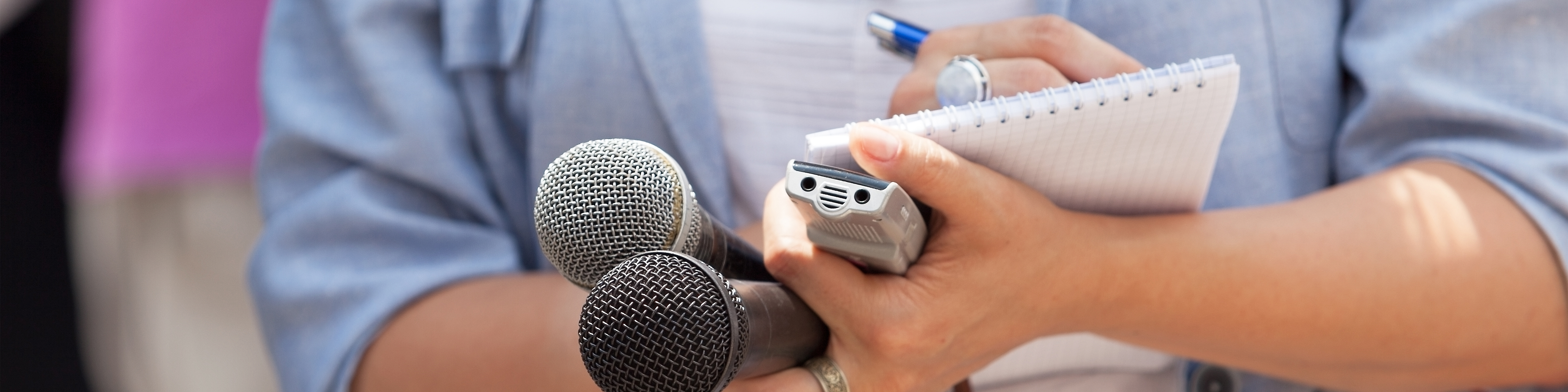 Photo of a reporter taking notes while holding two microphones and a voice recorder