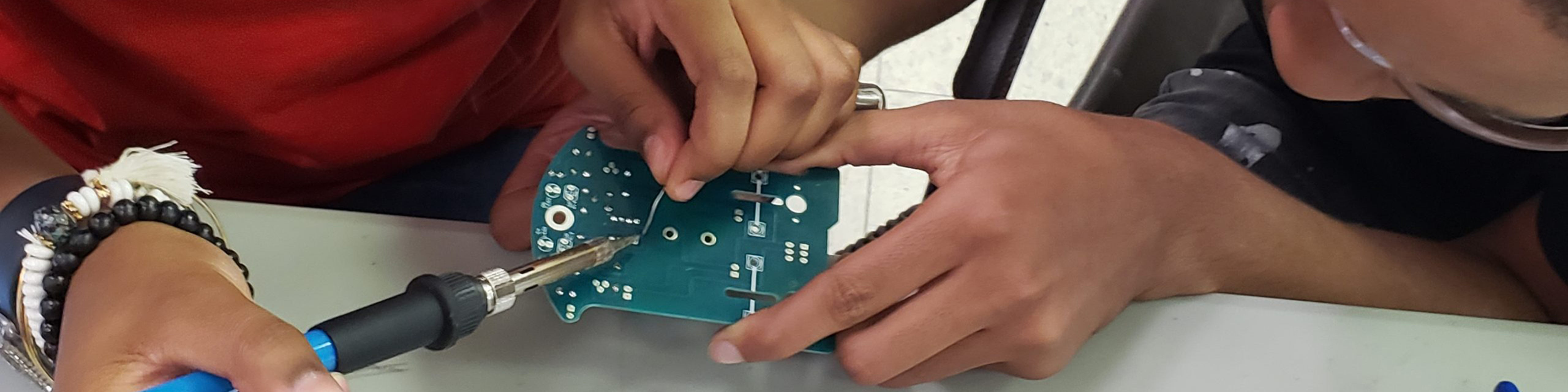Close up of two students' hands working to solder a circuit board