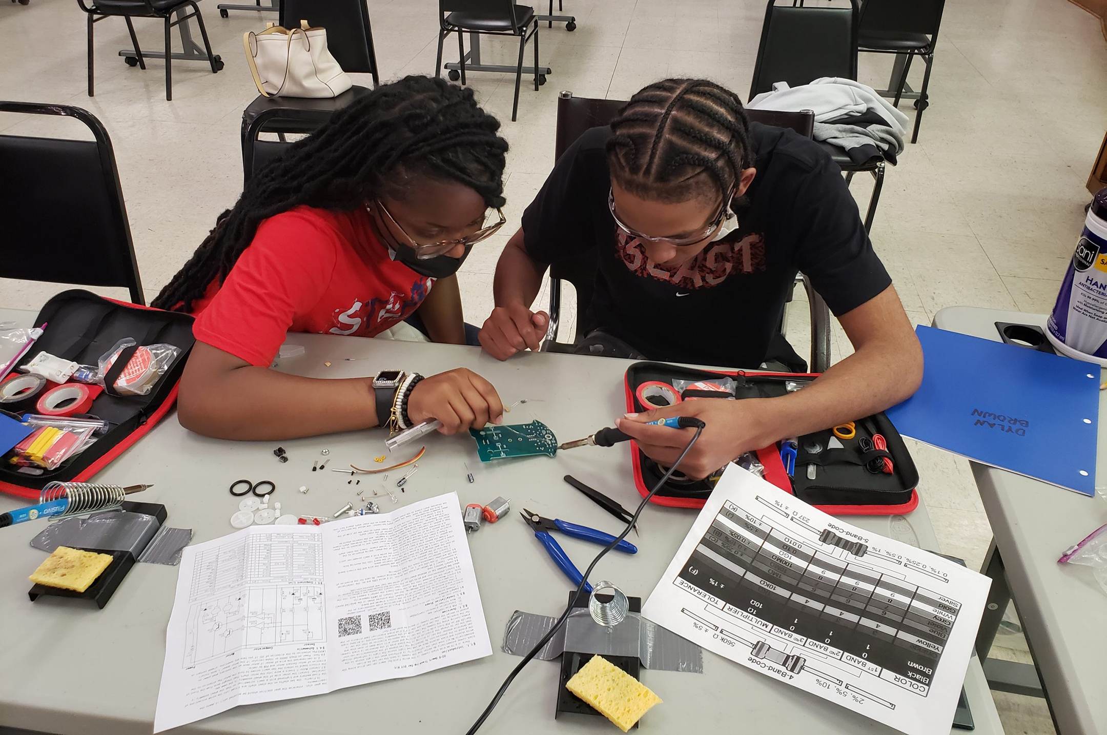 Two high school students sit at a desk covered with technical paperwork and tools while working on a circuitboard