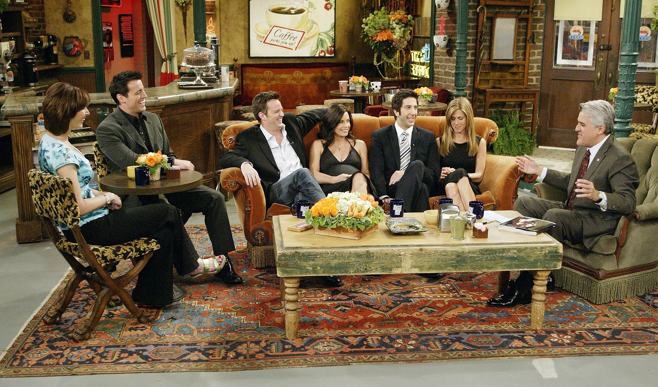 Seven people sit around a couch on a TV set talking