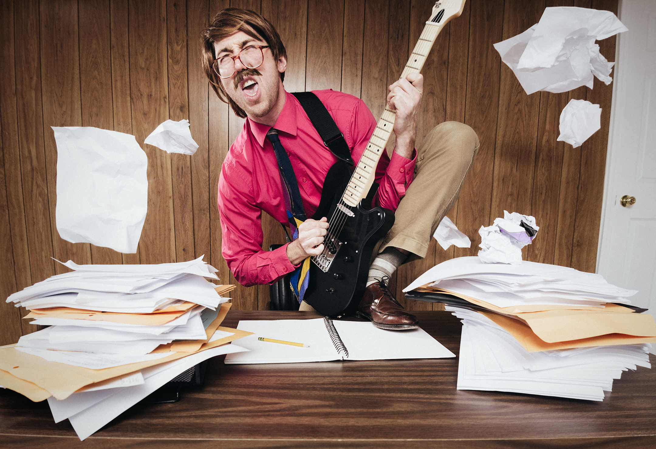 A white collar business man working in a retro 1980's style office stands on his desk piled with paperwork and documents, shredding some rock and roll on his electric guitar.