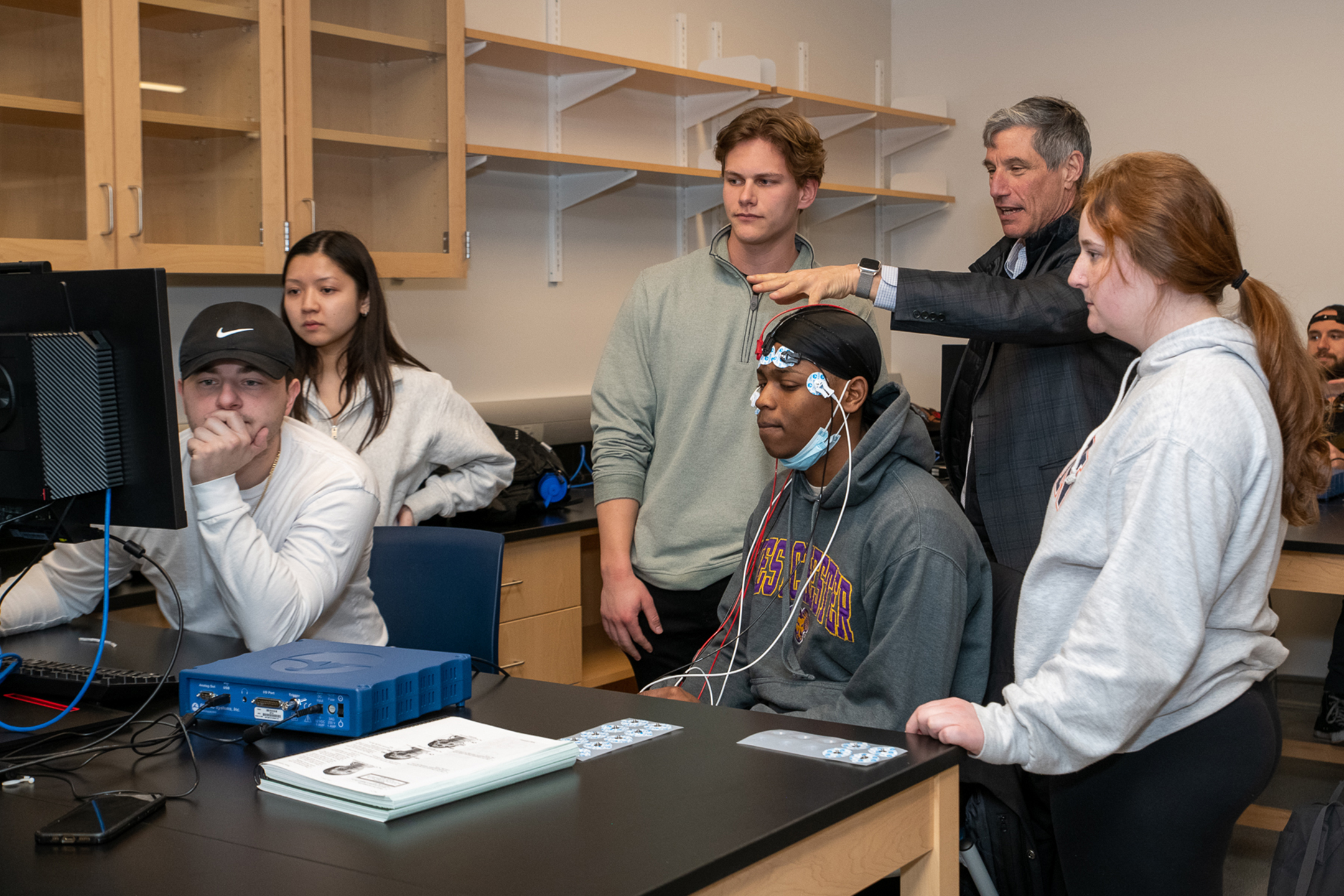 Dan Greenstein pointing at a computer  in a room with five other students while one sits at the monitor and another wears electrodes on his head