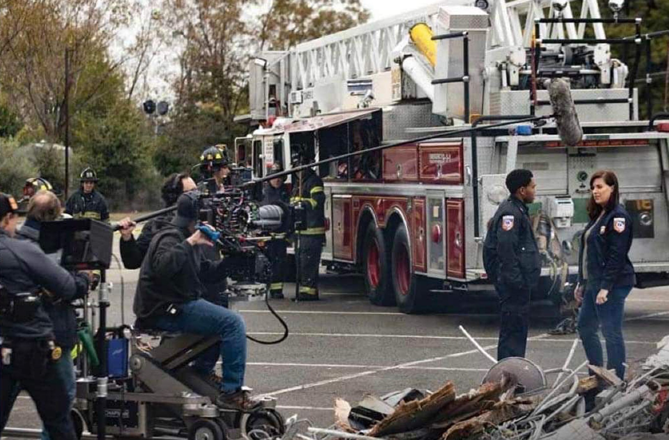 Two actors speak in front of a fire truck on a street while a crew films them on the left