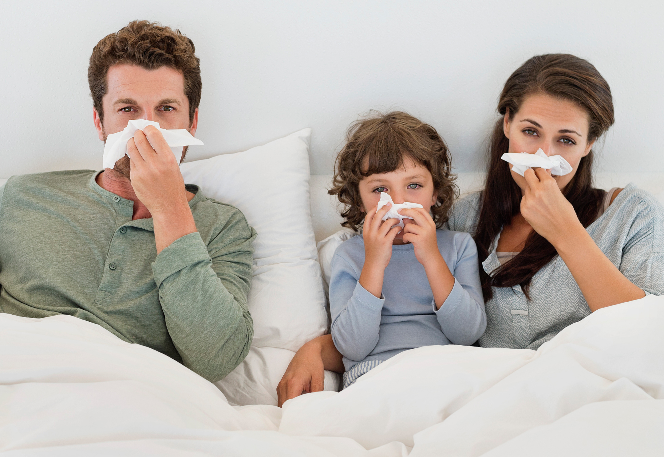 Two parents and a child in bed blowing their noses
