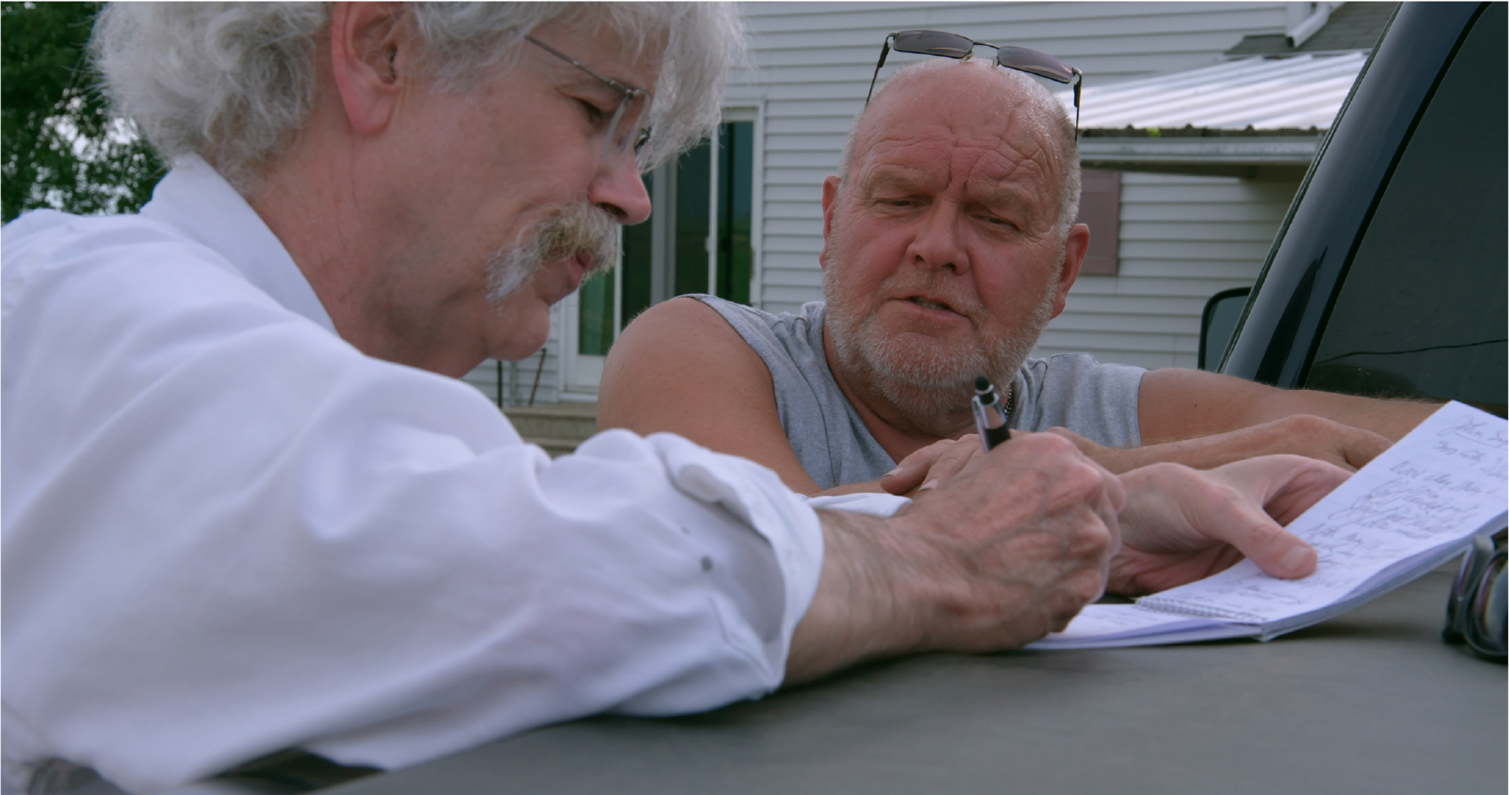 Man with mustache and glasses writes in a notebook as he talks to another man in a sleeveless t-shirt, both standing next to a pickup truck