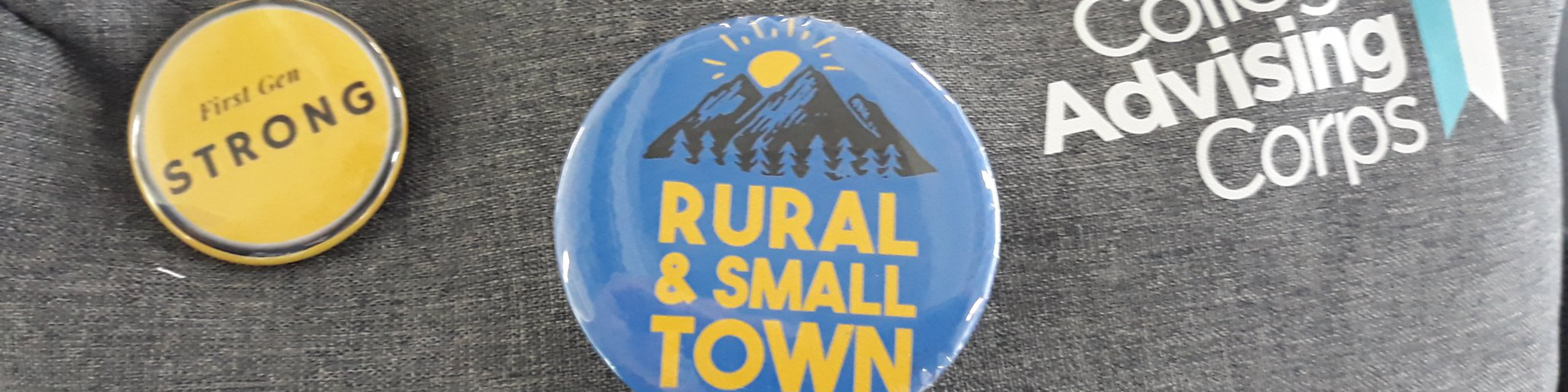 Closeup photo of a gray backpack with, from left, a round yellow pin that reads "first-gen strong," a round blue pin that reads "rural & small town," and the logo of College Advising Corps printed in white text