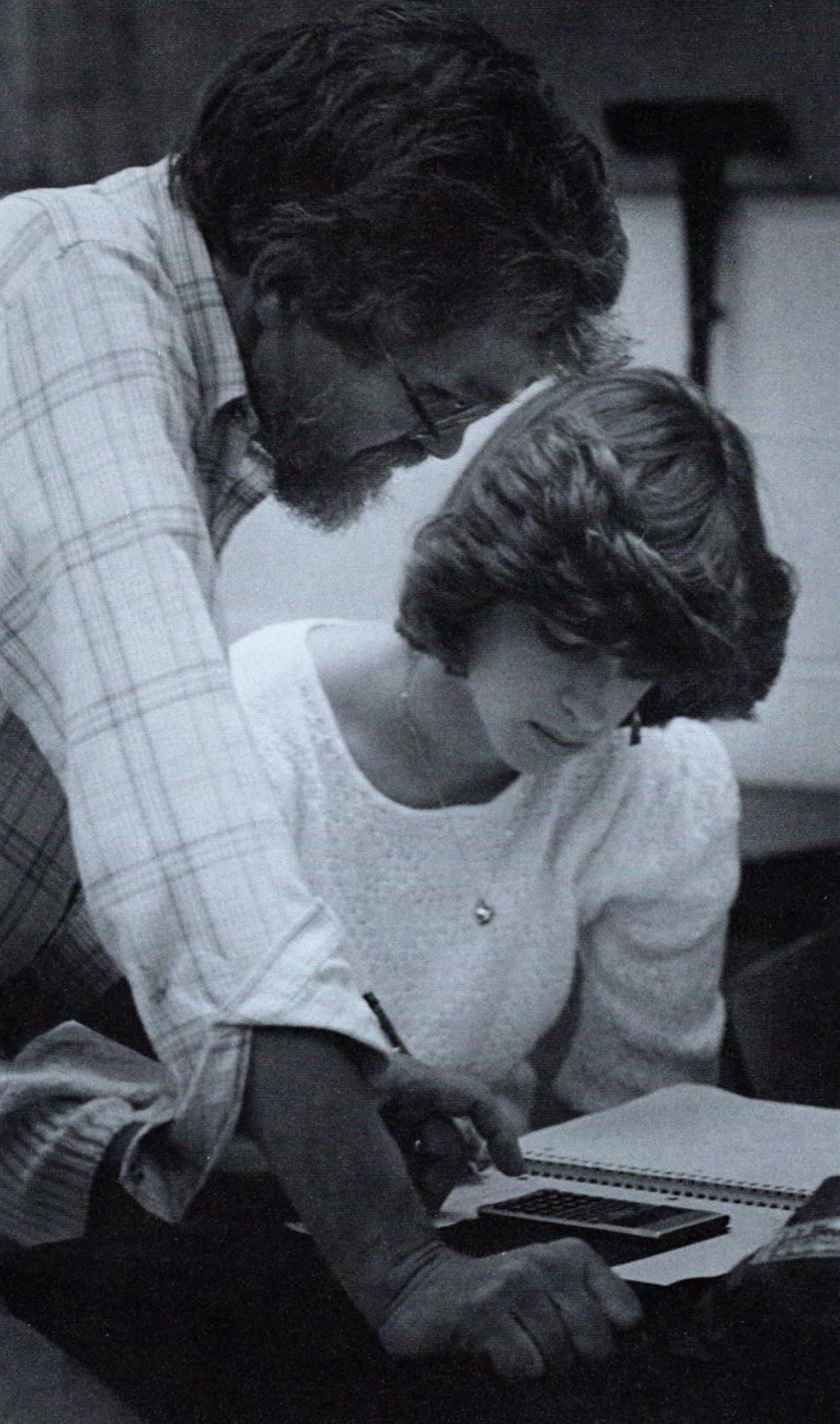 Black and white photo of a bearded male teacher, standing, working with a seated female college student who is using a notebook and calculator