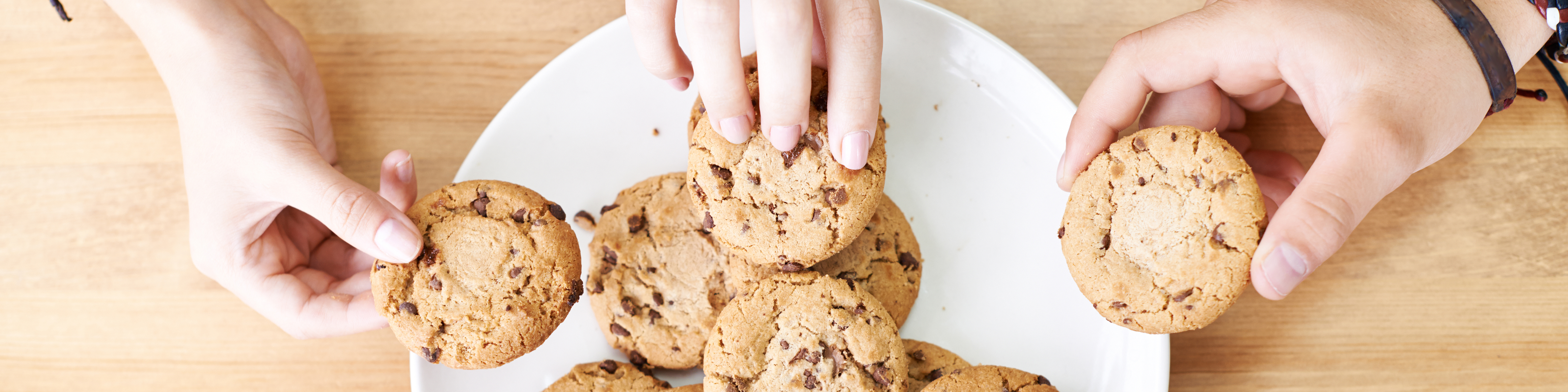  overhead photo of three hands reaching for a plate of chocolate chip cookies