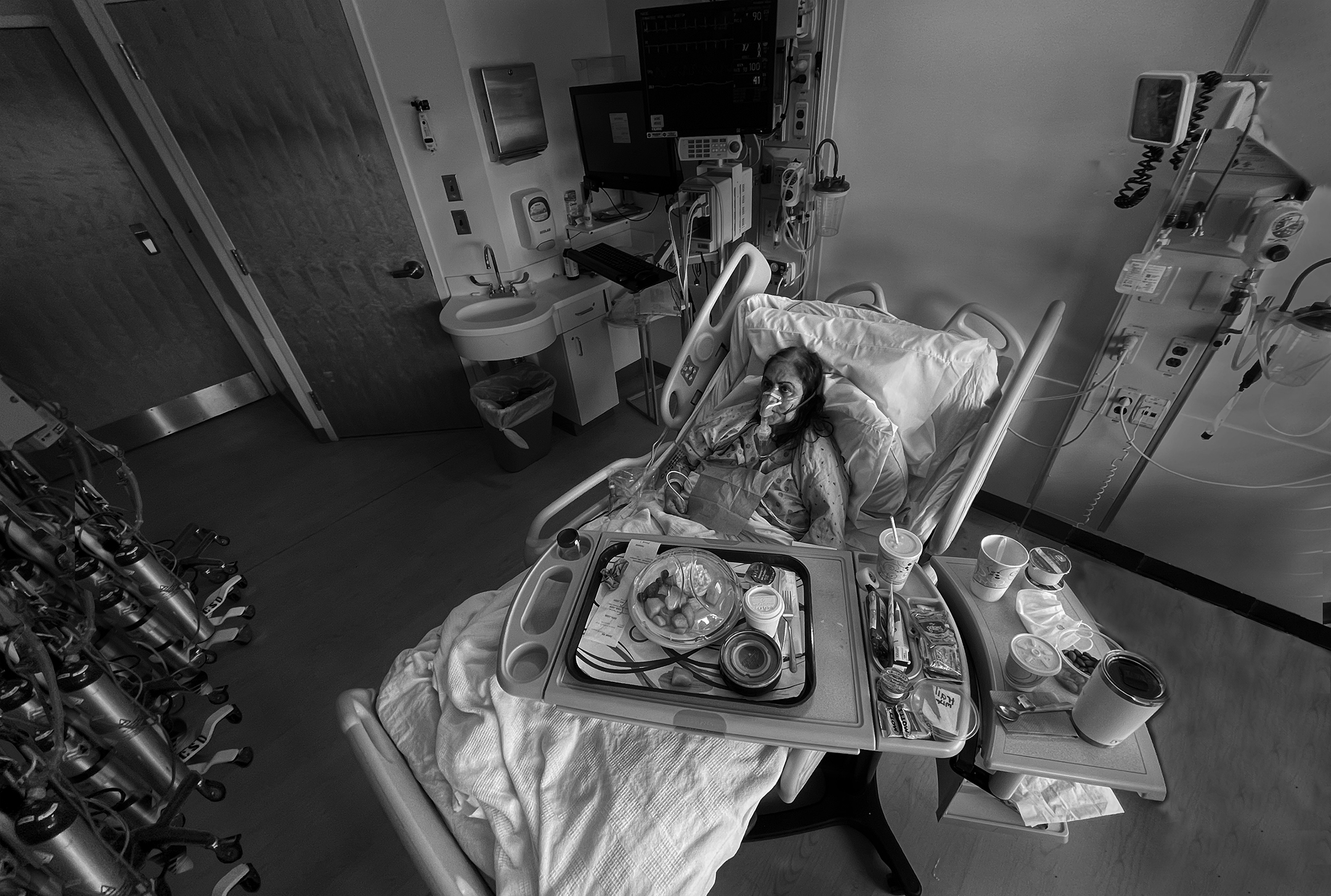 black and white photo of an old woman lying in a hospital bed, wearing an oxygen mask, and surrounded by food and hospital equipment