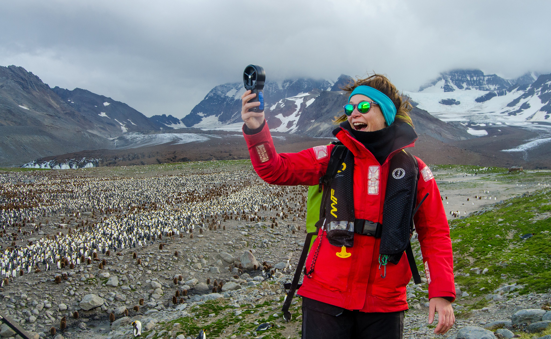smiling woman in cold weather gear smiling as she holds a small scientific instrument while standing above a herd of penguins