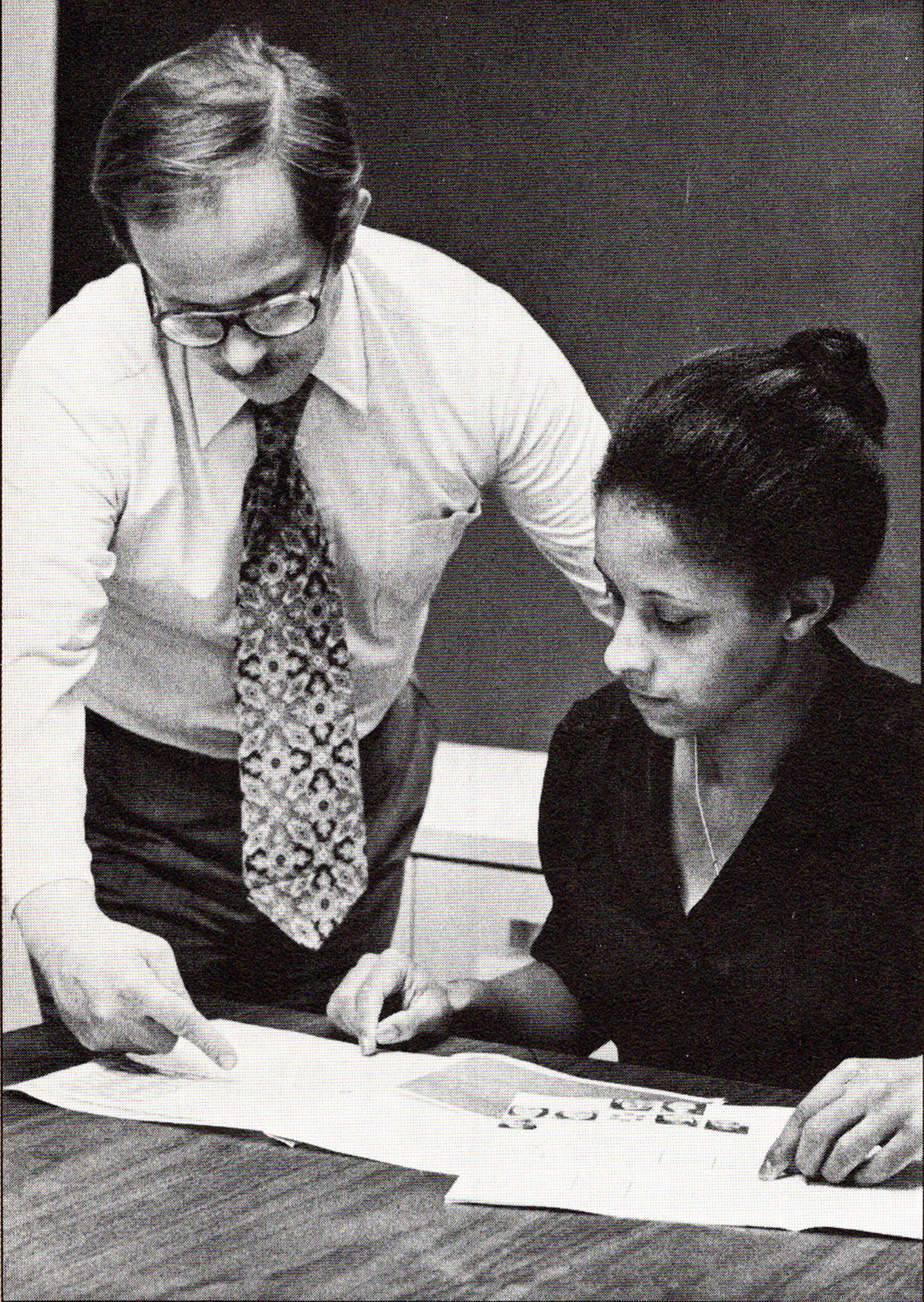 black and white photo of a male teacher standing while helping a seated female student with her career skills assessment