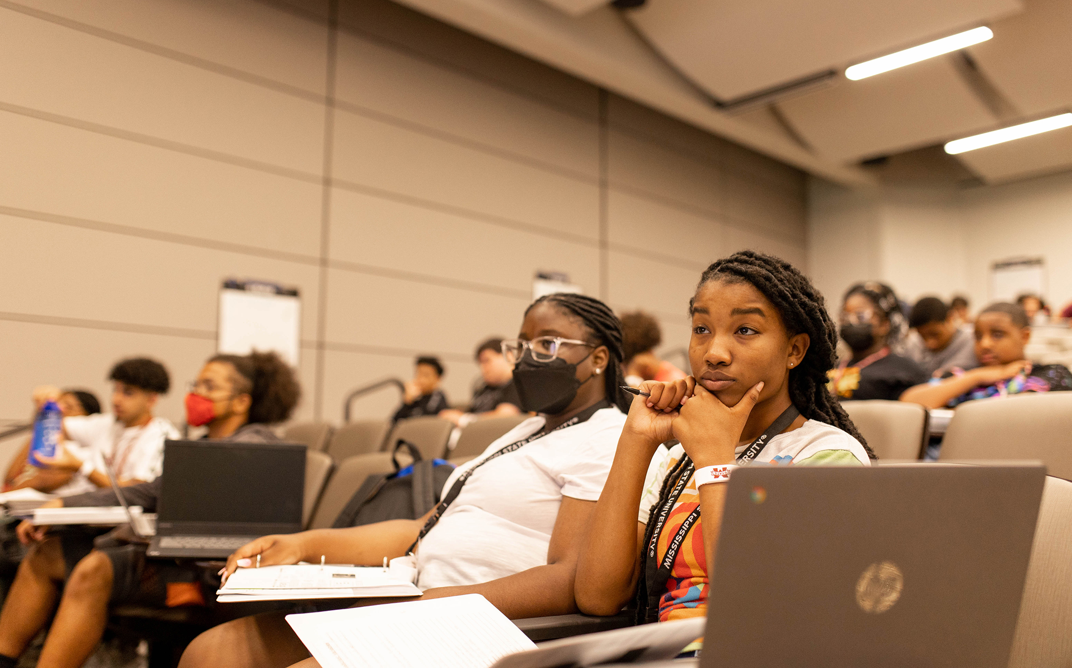 young black woman with her hand on her chin listens intently in a lecture hall, a notebook and laptop open in front of her