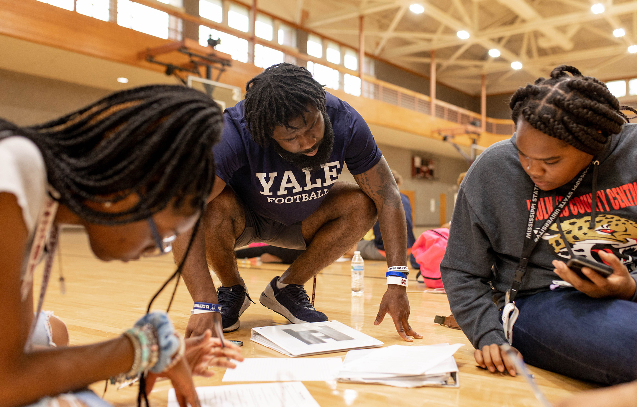 a black male student teaching assistant in a yale t-shirt works with two young black women on a project on a gym floor