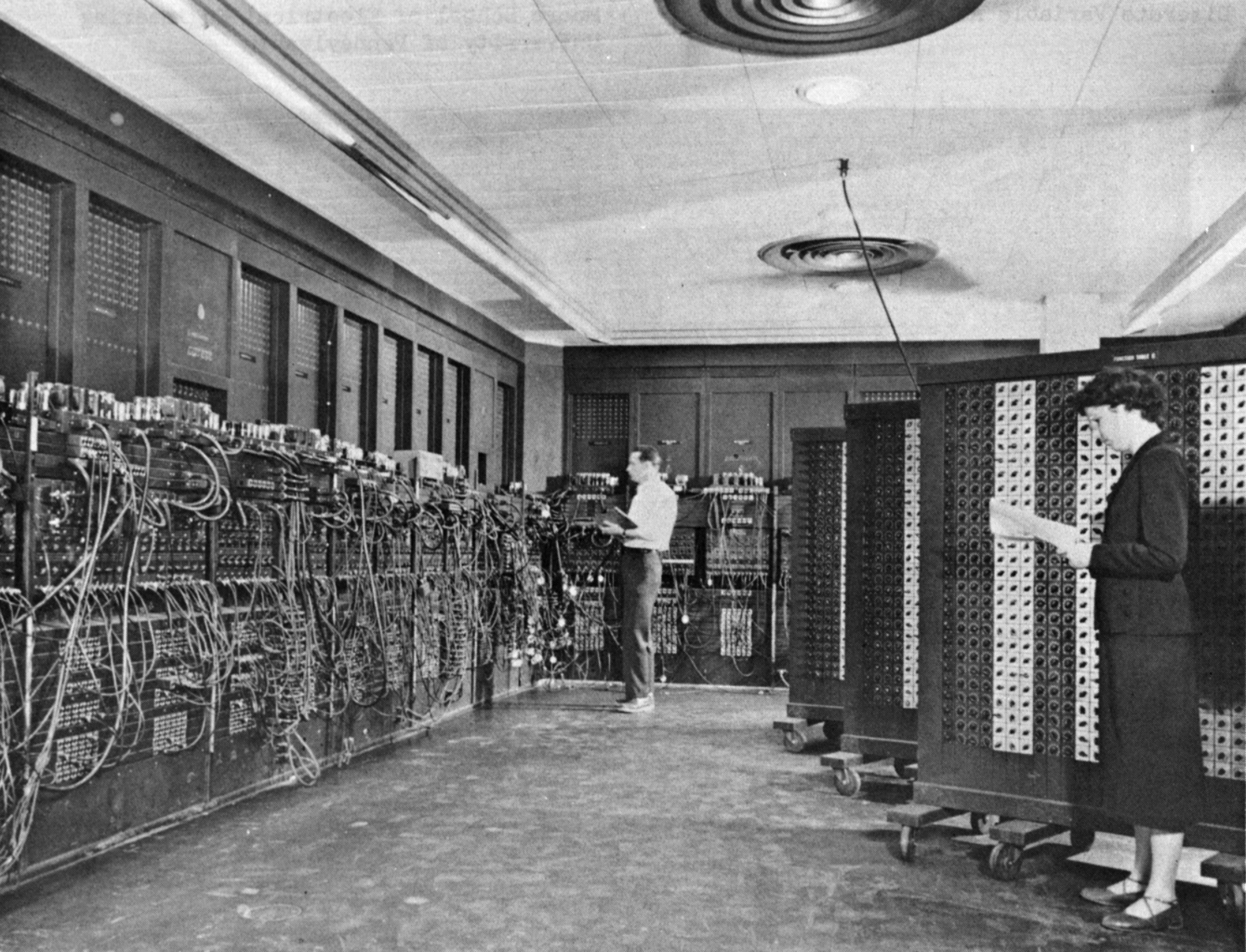 Black and white photo of a woman and man in room working on the room-sized eniac computer