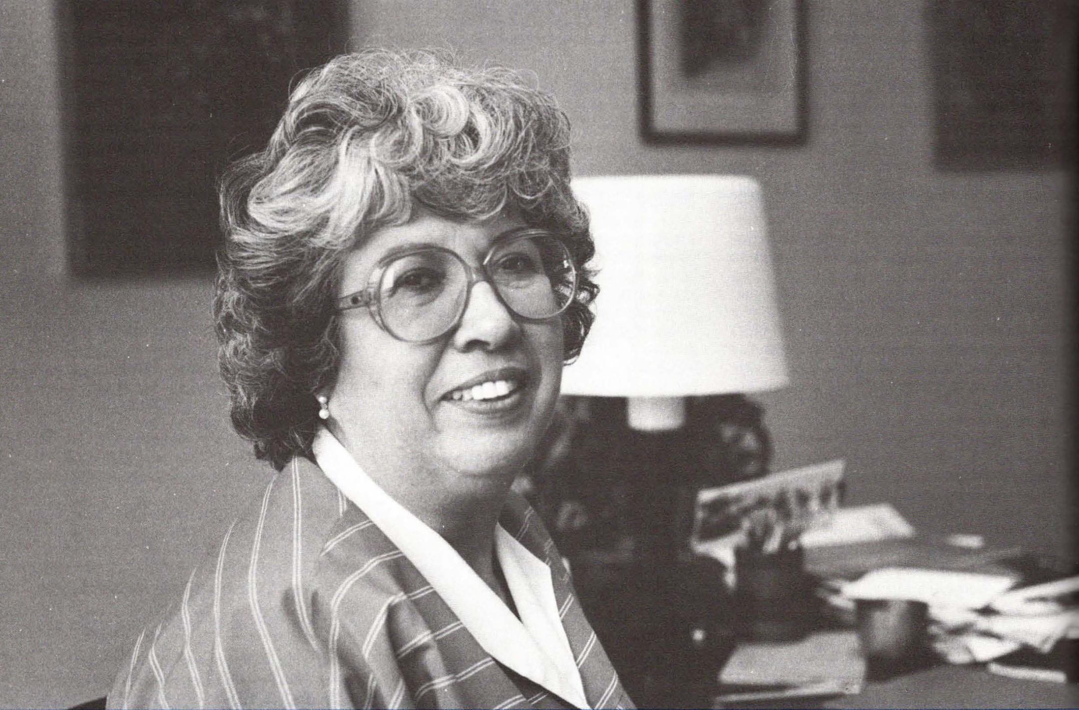 Black and white photo of a woman wearing glasses, smiling as she sits at her desk and looks at the camera