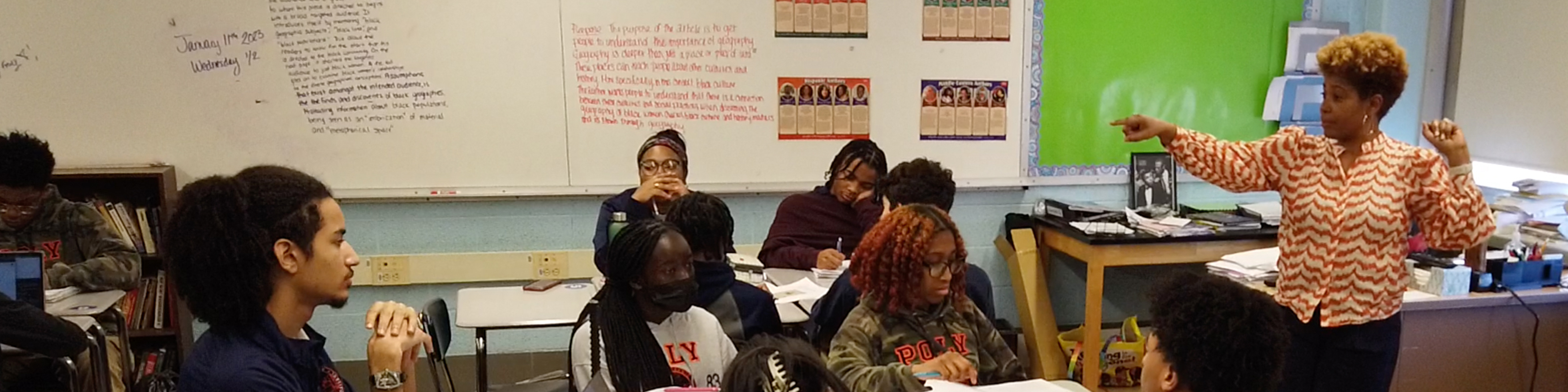 black female teacher, standing at right, points while discussing a lesson in her classroom of seated students