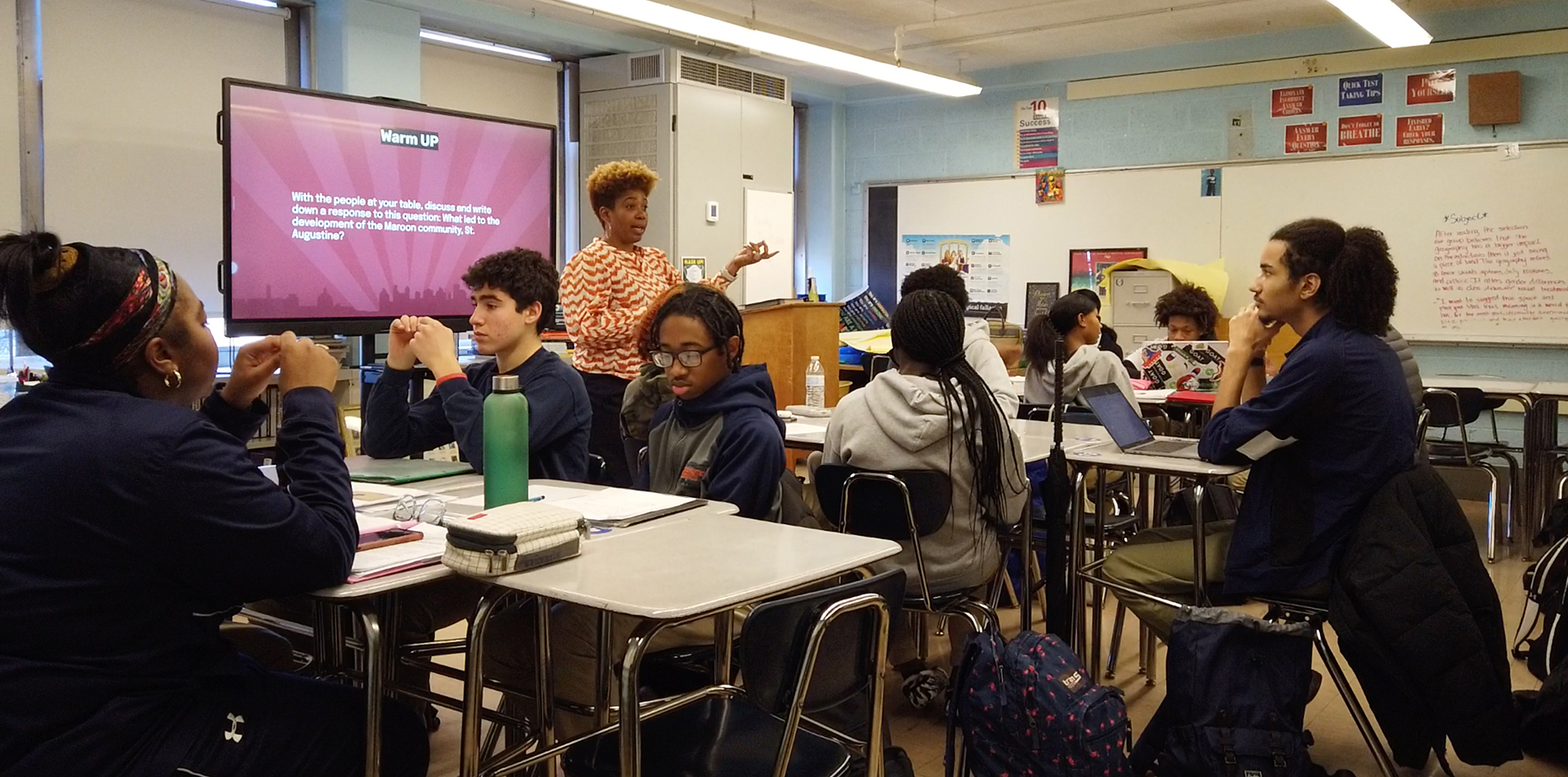 black female teacher, standing at left, gestures while discussing a lesson in her classroom of seated students