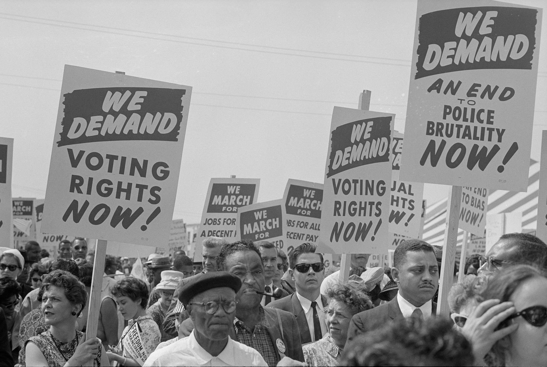 black and white photo of a crowd of black and white marchers carrying signs for equal rights, integrated schools, decent housing, and an end to bias