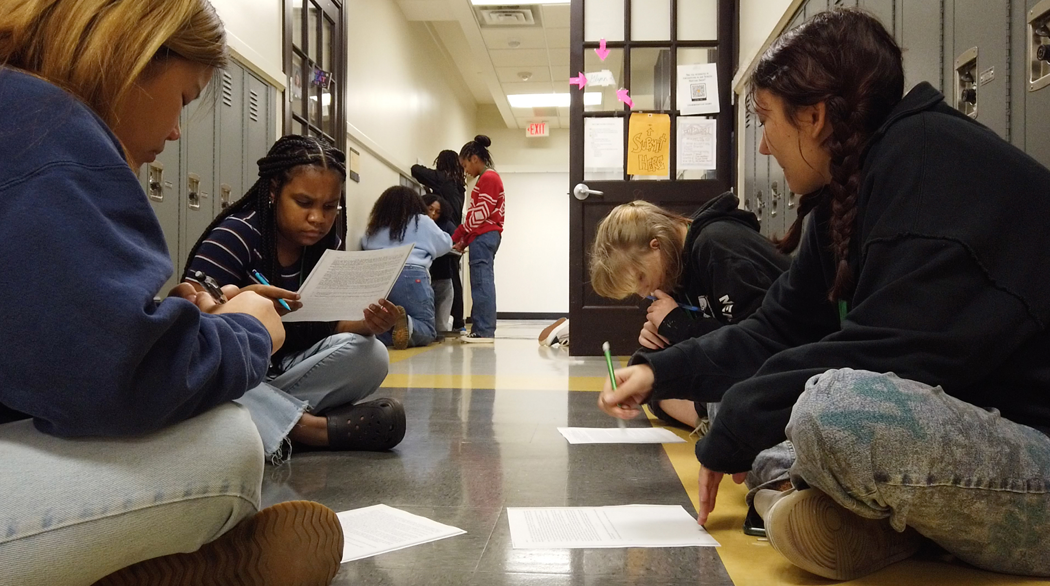 Four students, all female, sit in a high school hallway and work on a project with pencil and paper