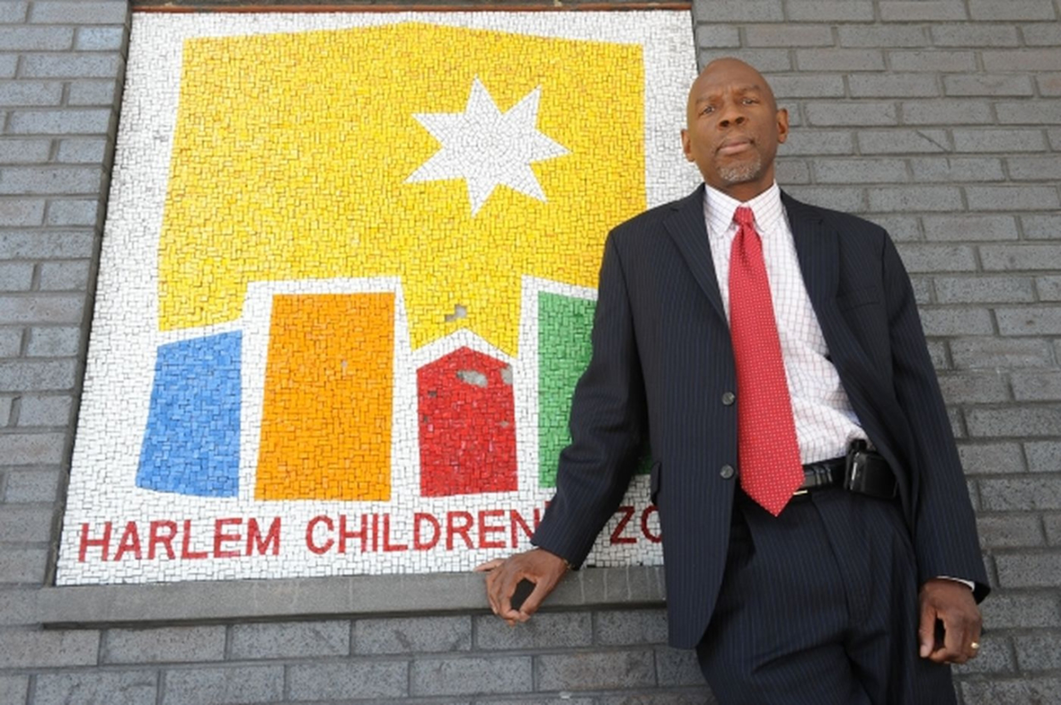 Geoffrey Canada standing against a wall with a mosaic logo in yellow, blue, orange, red, and green for Harlem Children's Zone
