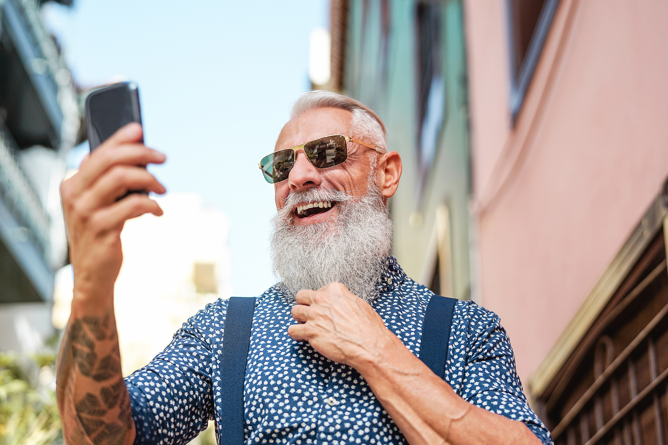 an older man with a long beard and wearing sunglasses smiles as he takes a selfie with his smartphone