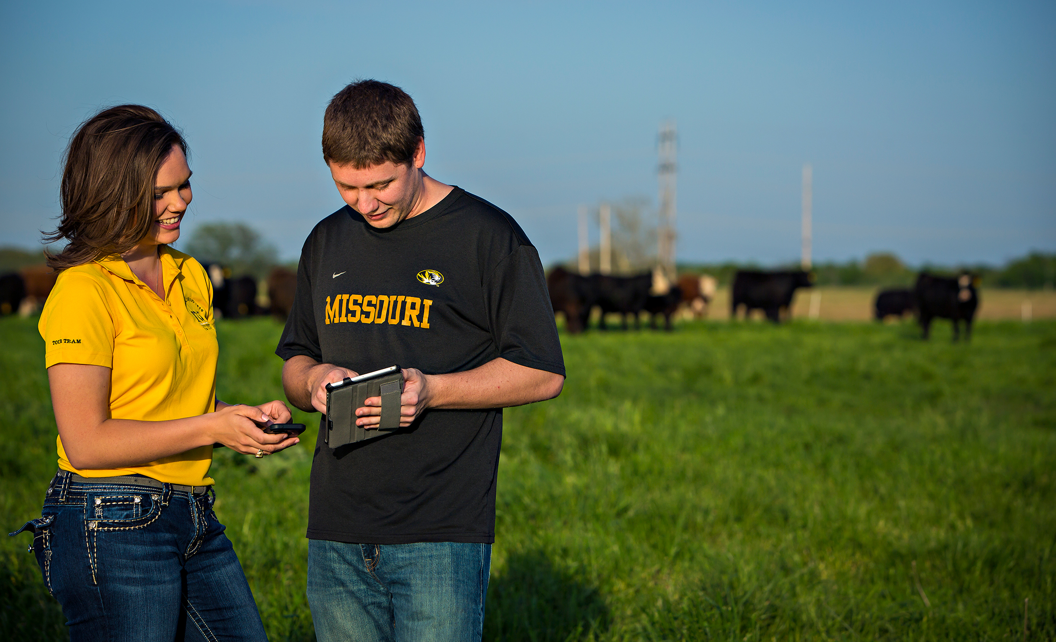 Two students, one female in a yellow polo shirt on the left and the other male in a black t-shirt on the right, smile as they look at a tablet computer. A herd of cows is in the background.