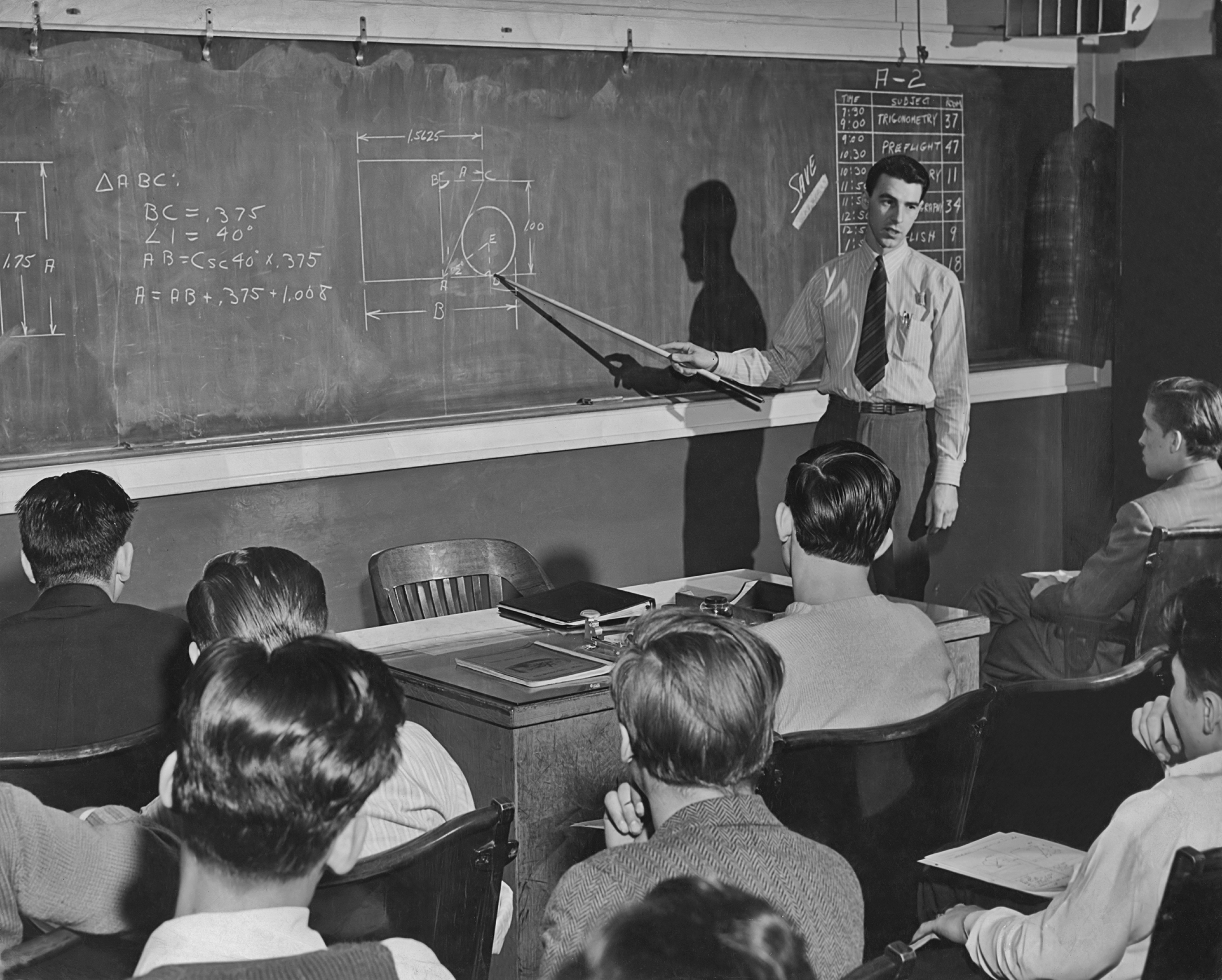 Black and white photo of a male teacher in front of a blackboard pointing at an equation with a stick.