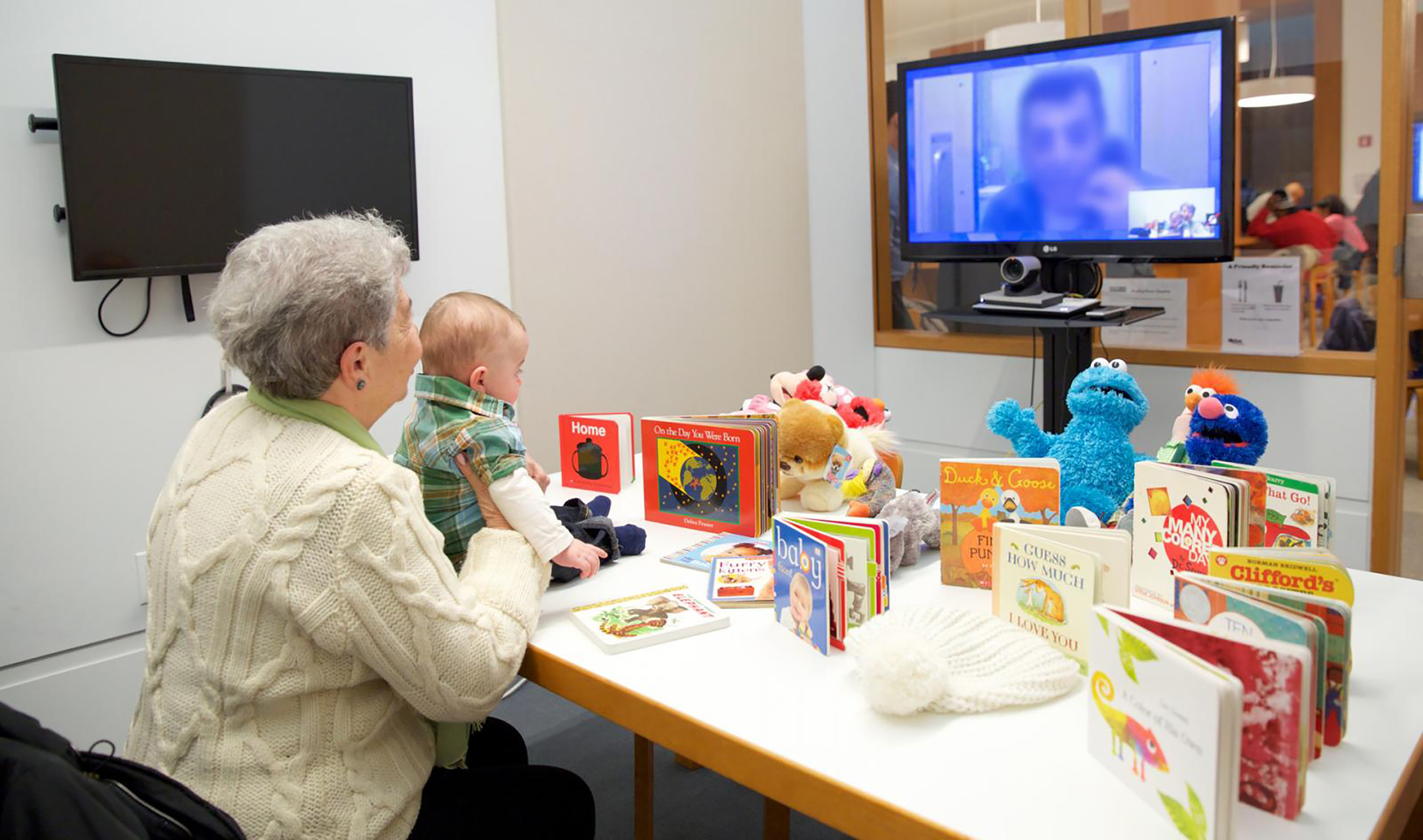 Elderly woman holding a small child sit at a table covered in toys and kids books while connecting with a male loved one, who's face is obscured, via a video screen