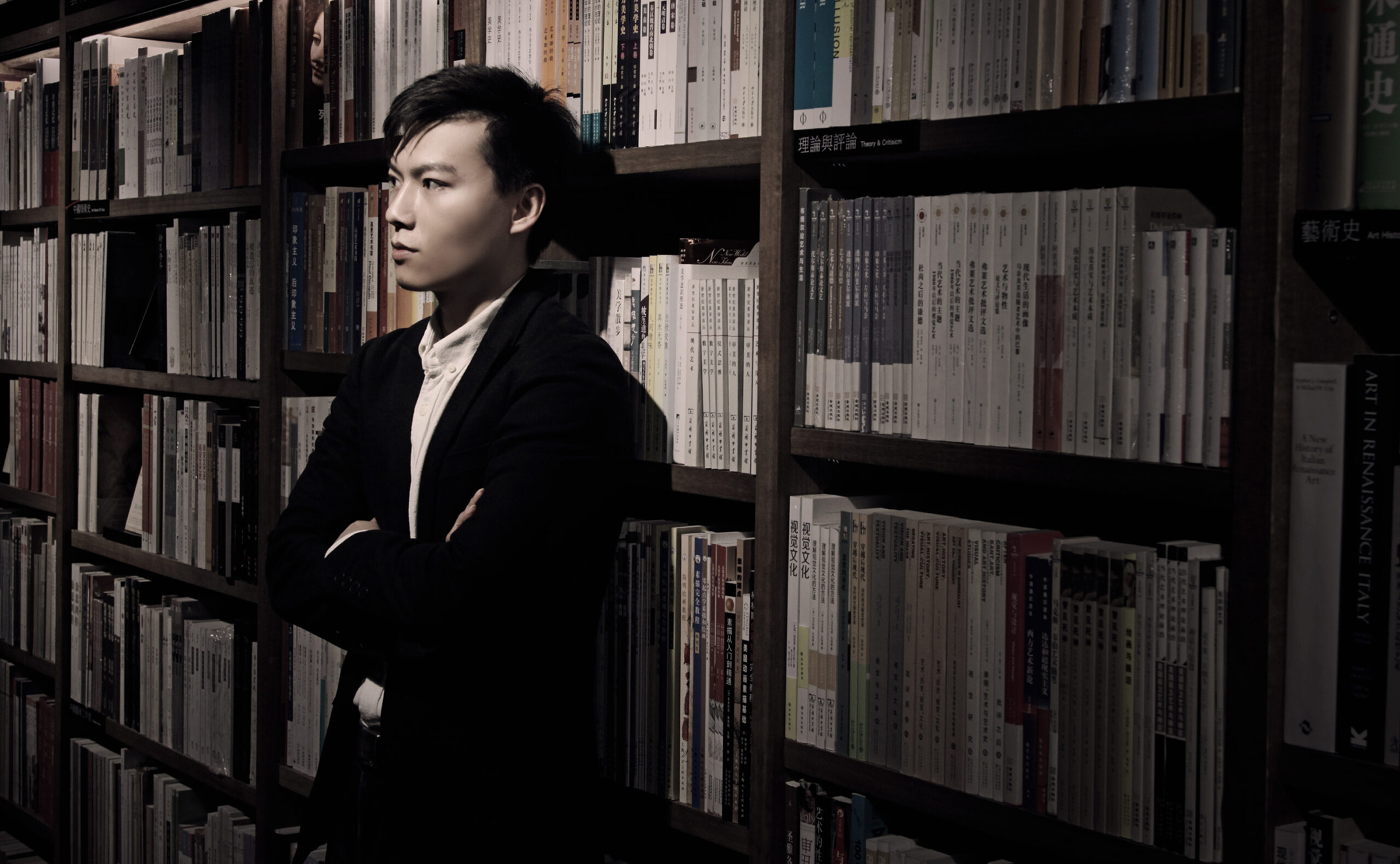 Photo of a young Chinese man leaning pensively against a library shelf of books