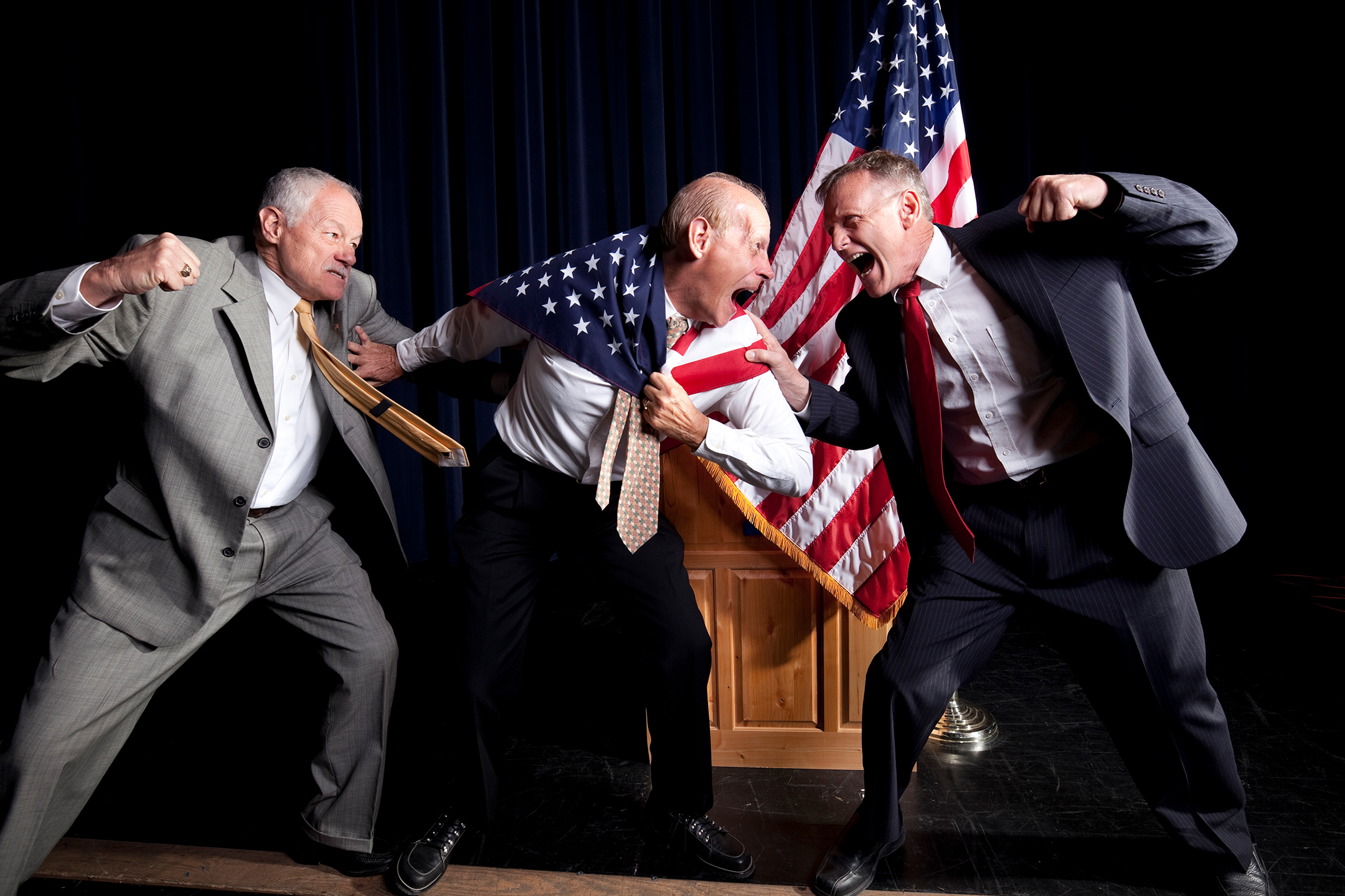Three old white men, two on the left, one on the right, about to throw punches at each other while on a stage. The man in the middle is wrapped in an American flag.