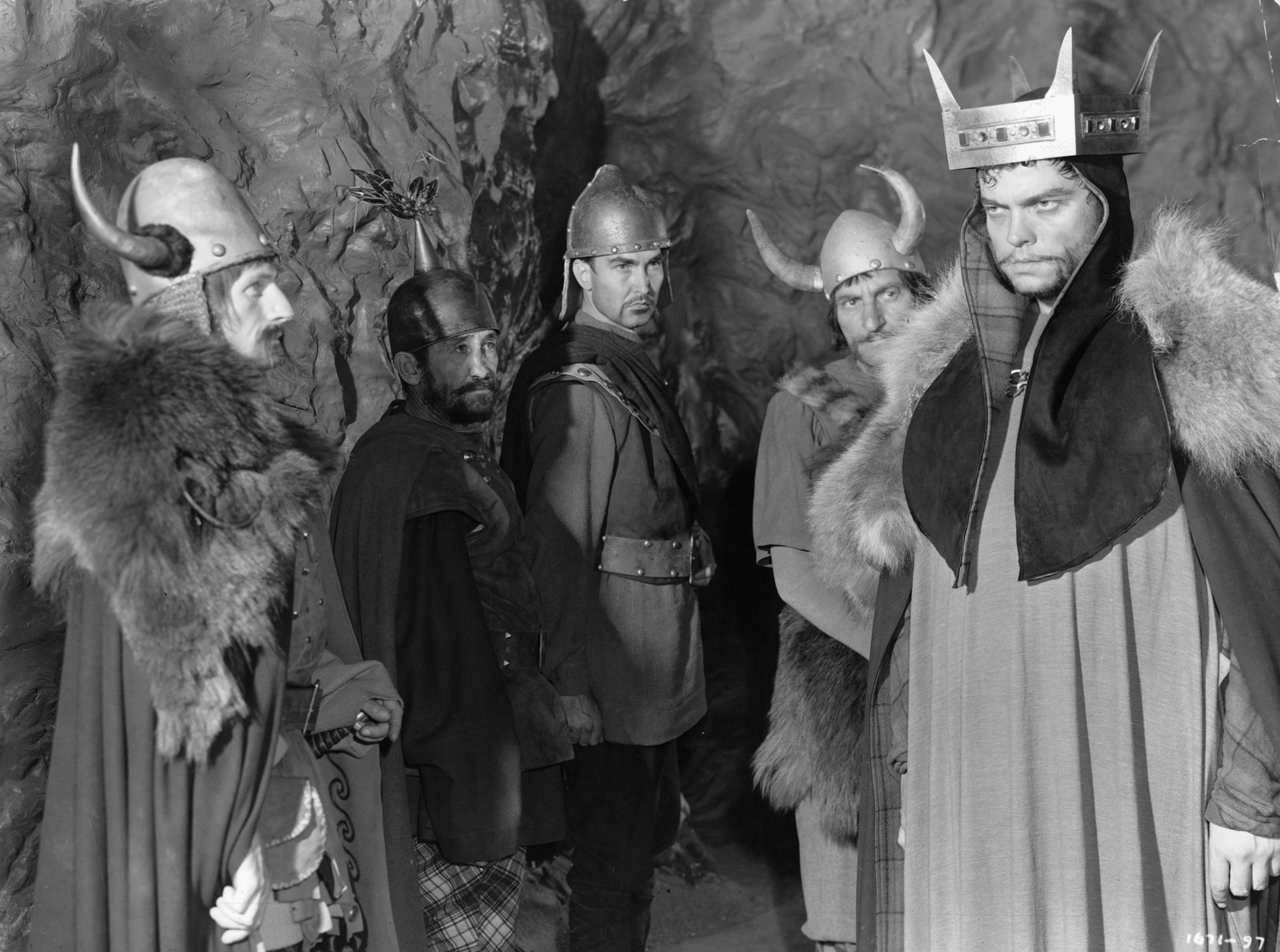 Orson Welles, wearing a crown and robes, looks into the distance while being watched by men in Viking uniforms in a scene from the film 'Macbeth'