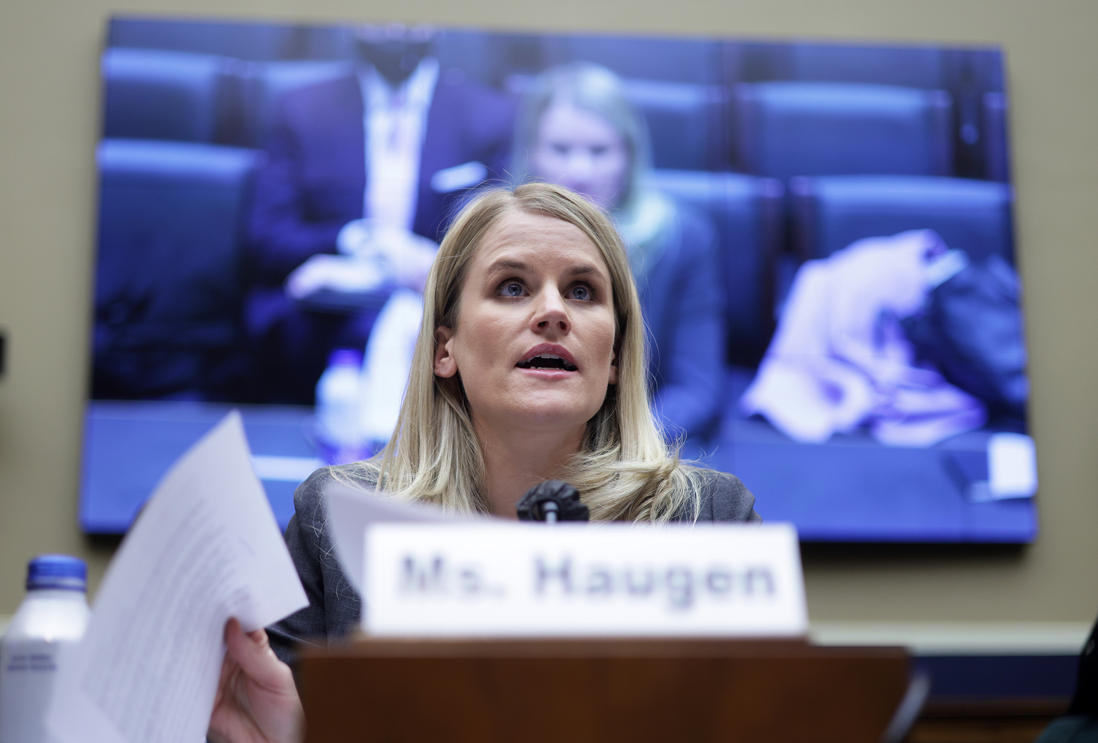 Woman speaking at desk with a television behind her showing her speaking